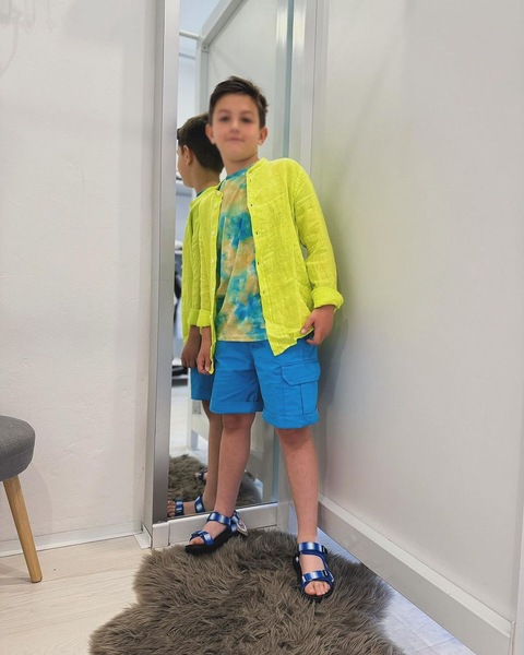 Un ragazzo in un negozio di abbigliamento prova un vestito: maglione giallo, pantaloncini blu, sandali. In piedi davanti a uno specchio.