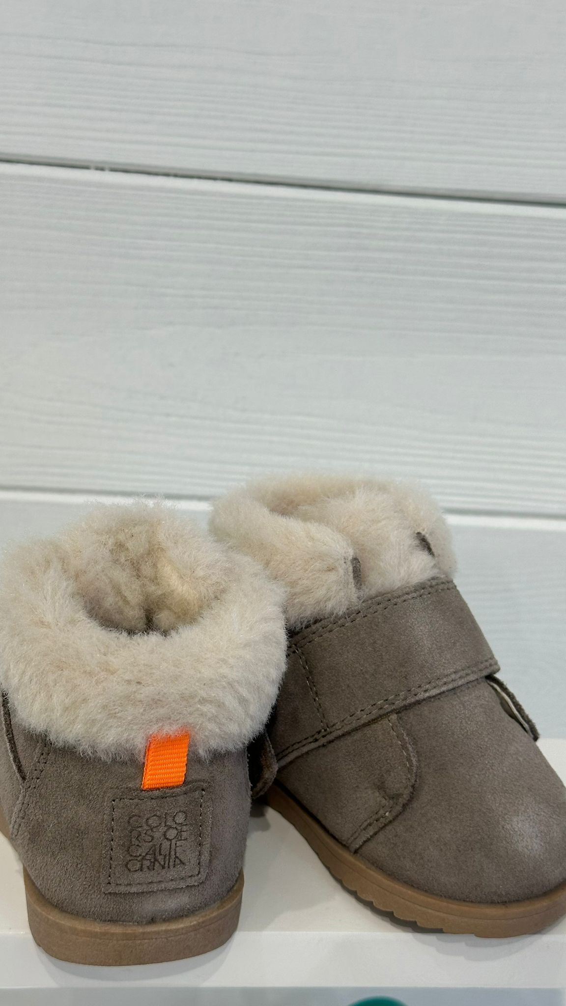 Stivali grigi per bambini con fodera pelosa e cinturino in velcro, su uno scaffale bianco.