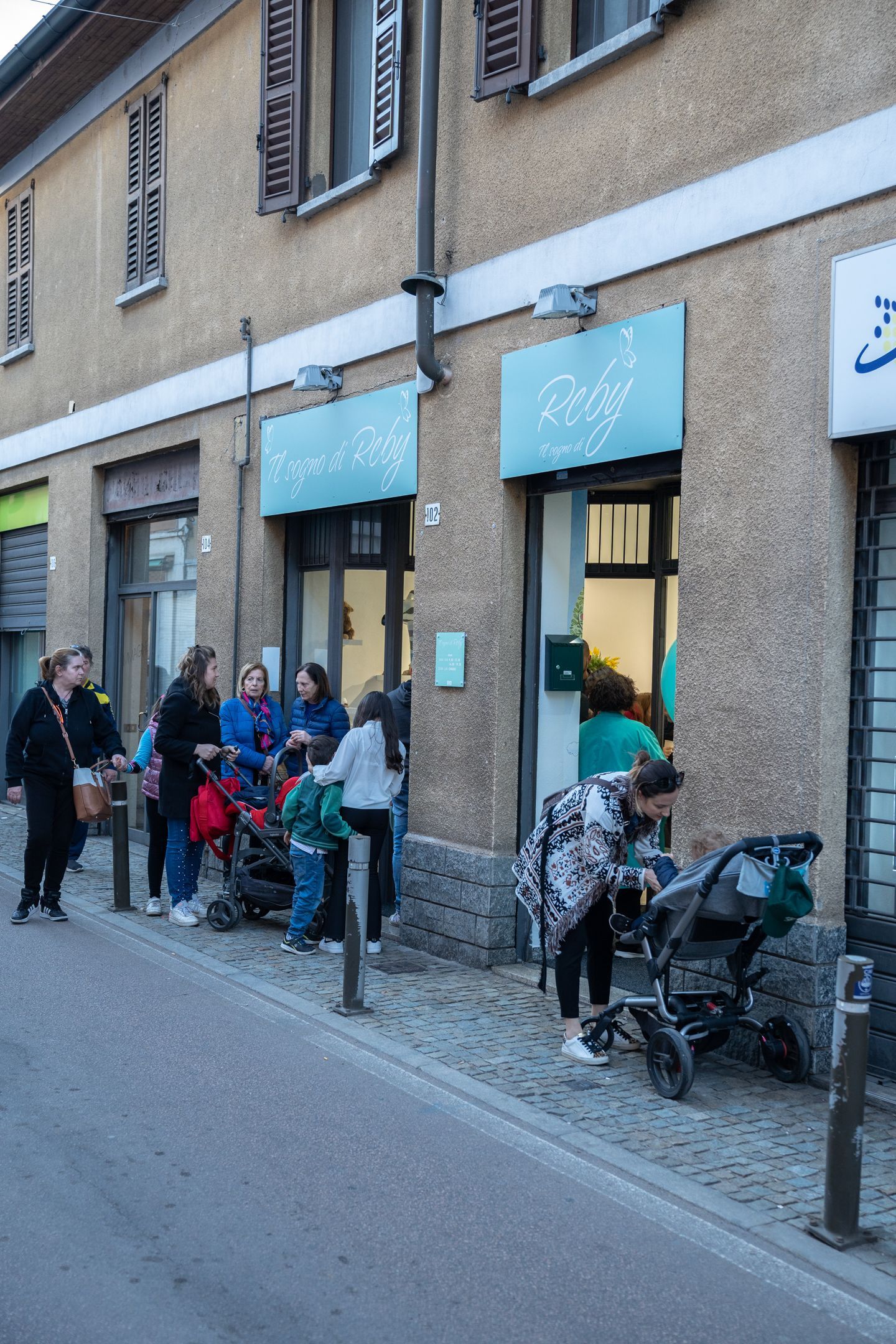Persone in fila fuori da un edificio con cartelli blu. Sono presenti diversi passeggini.