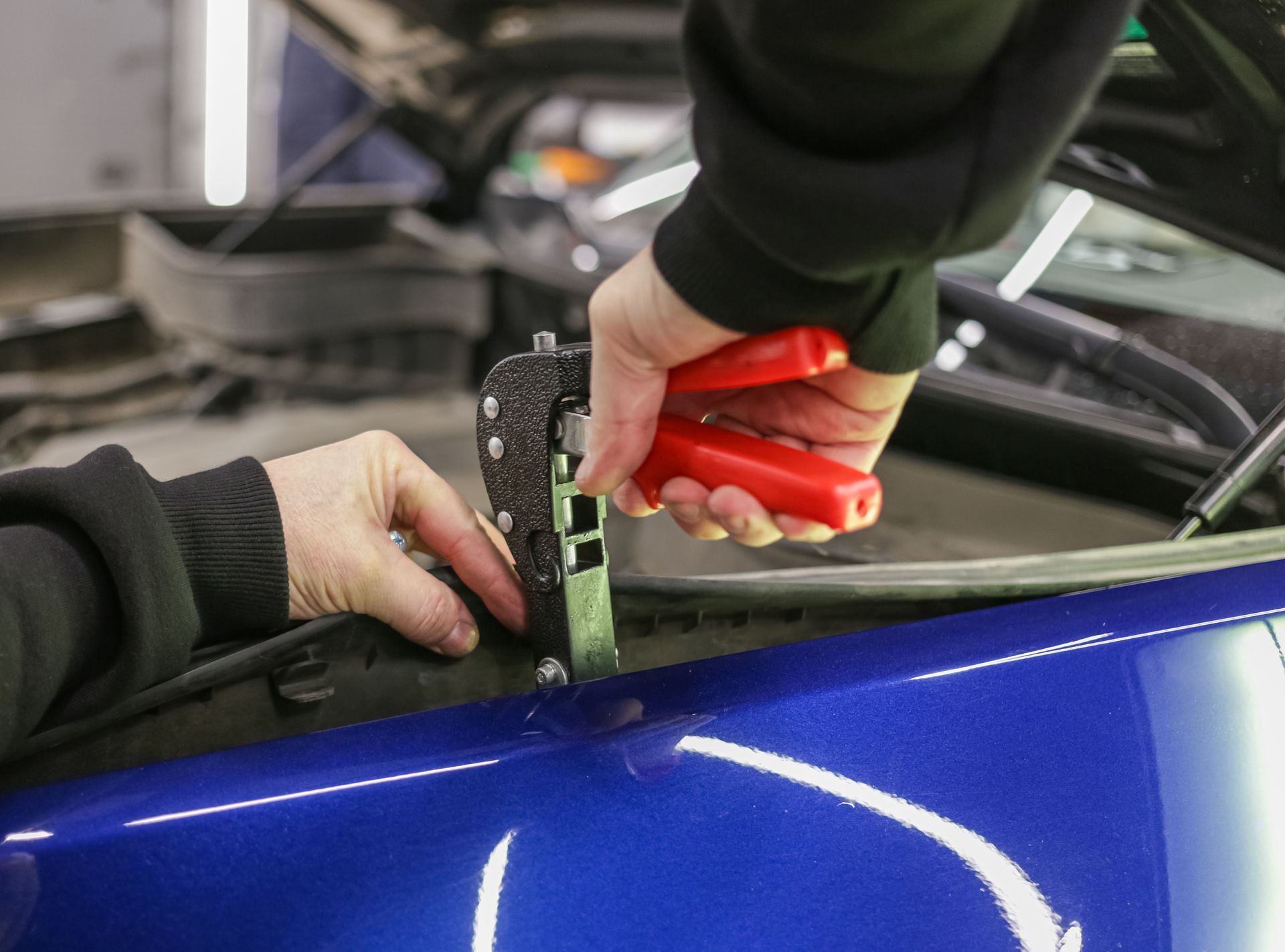 Hands using a red tool to pry a blue car door panel open in a garage