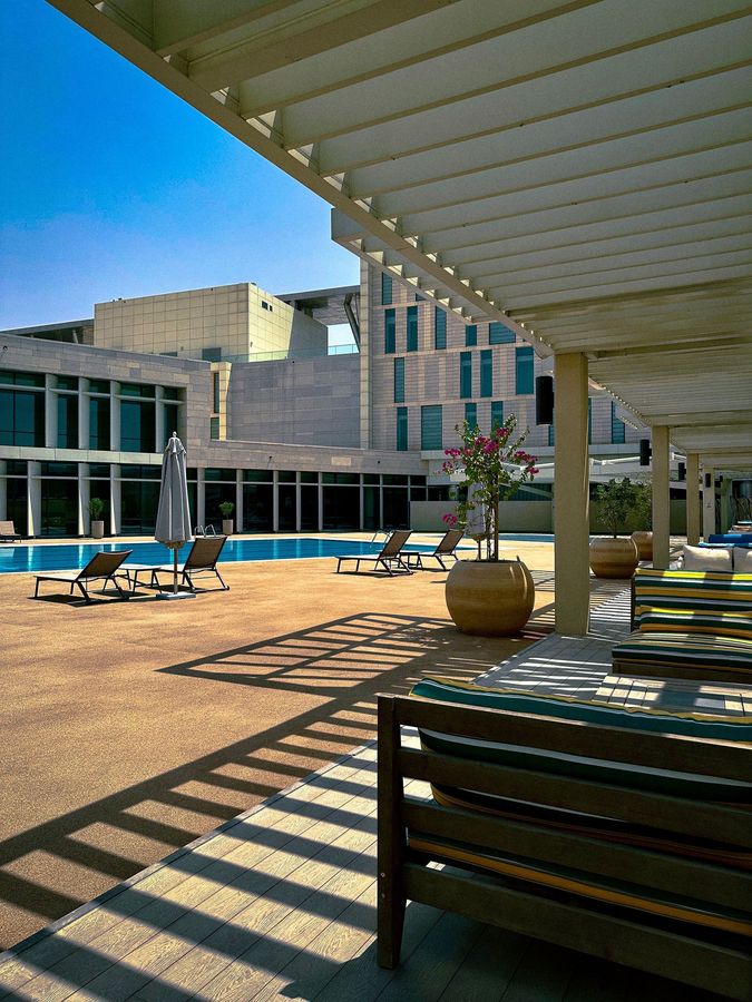 Modern hotel courtyard with lounge chairs, pergola shadows, and a pool under a clear blue sky