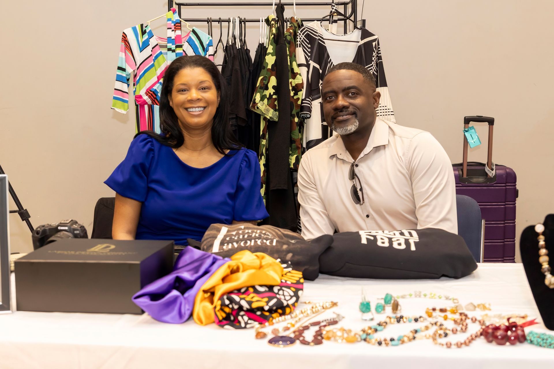 Two people at a table displaying colorful shirts, beaded jewelry, and a black tote in a vendor booth