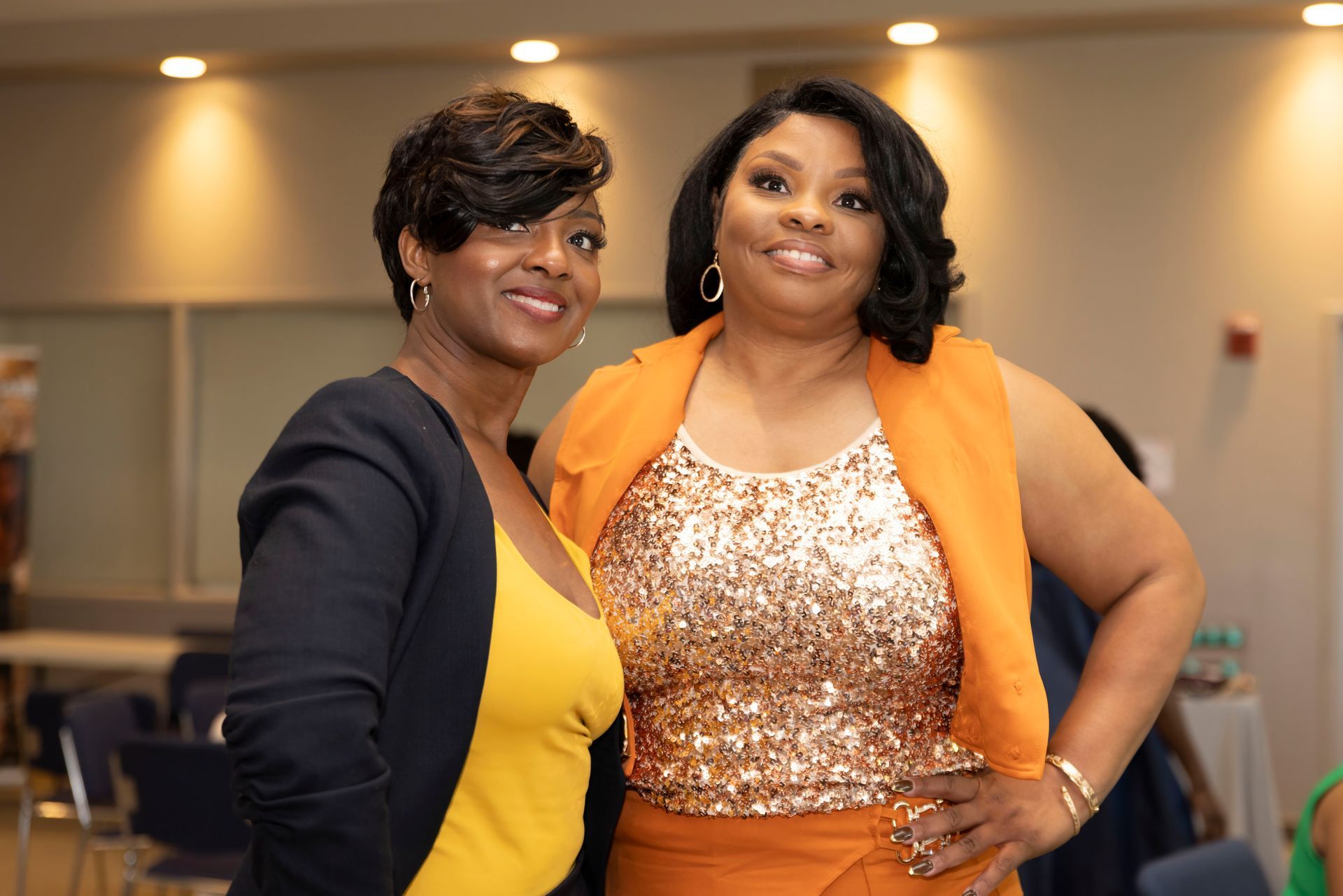 Two women smiling and posing together indoors, one in yellow and navy, the other in a sequined top and orange shawl