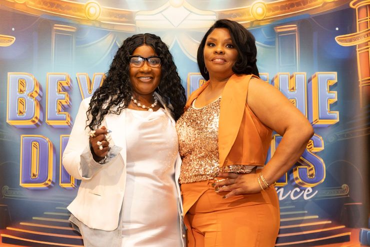 Two women posing at a blue-and-gold event backdrop, one holding a microphone and smiling.