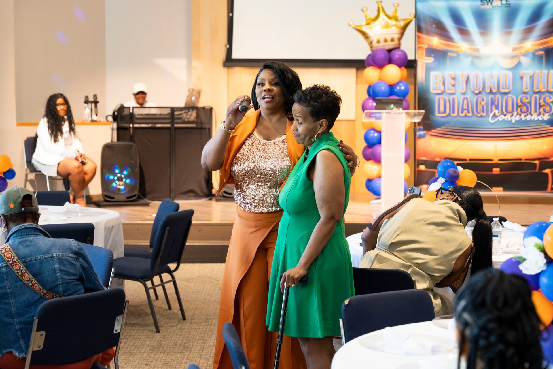 Two women posing at a banquet hall event with blue chairs and a balloon backdrop reading “Beyond the Diamond”