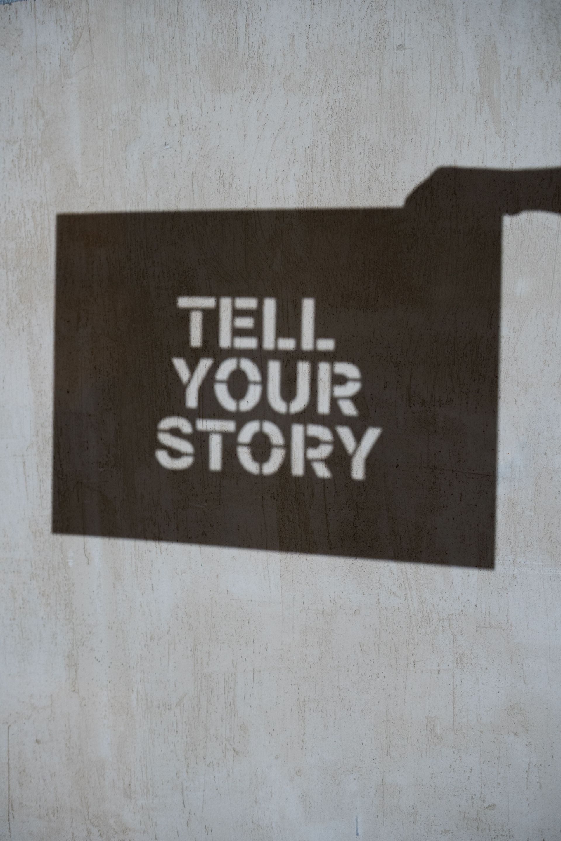 Black sign with white stencil text reading “TELL YOUR STORY” on a light wall