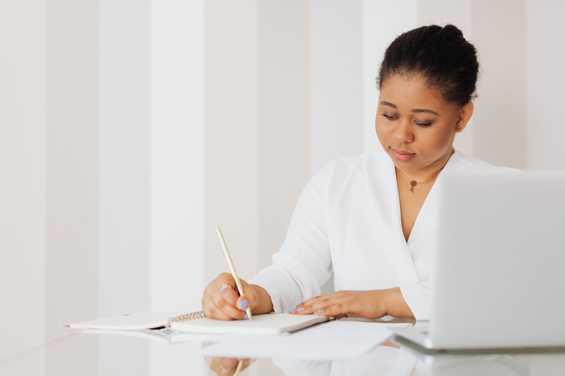 Person writing in a notebook beside a laptop at a white desk