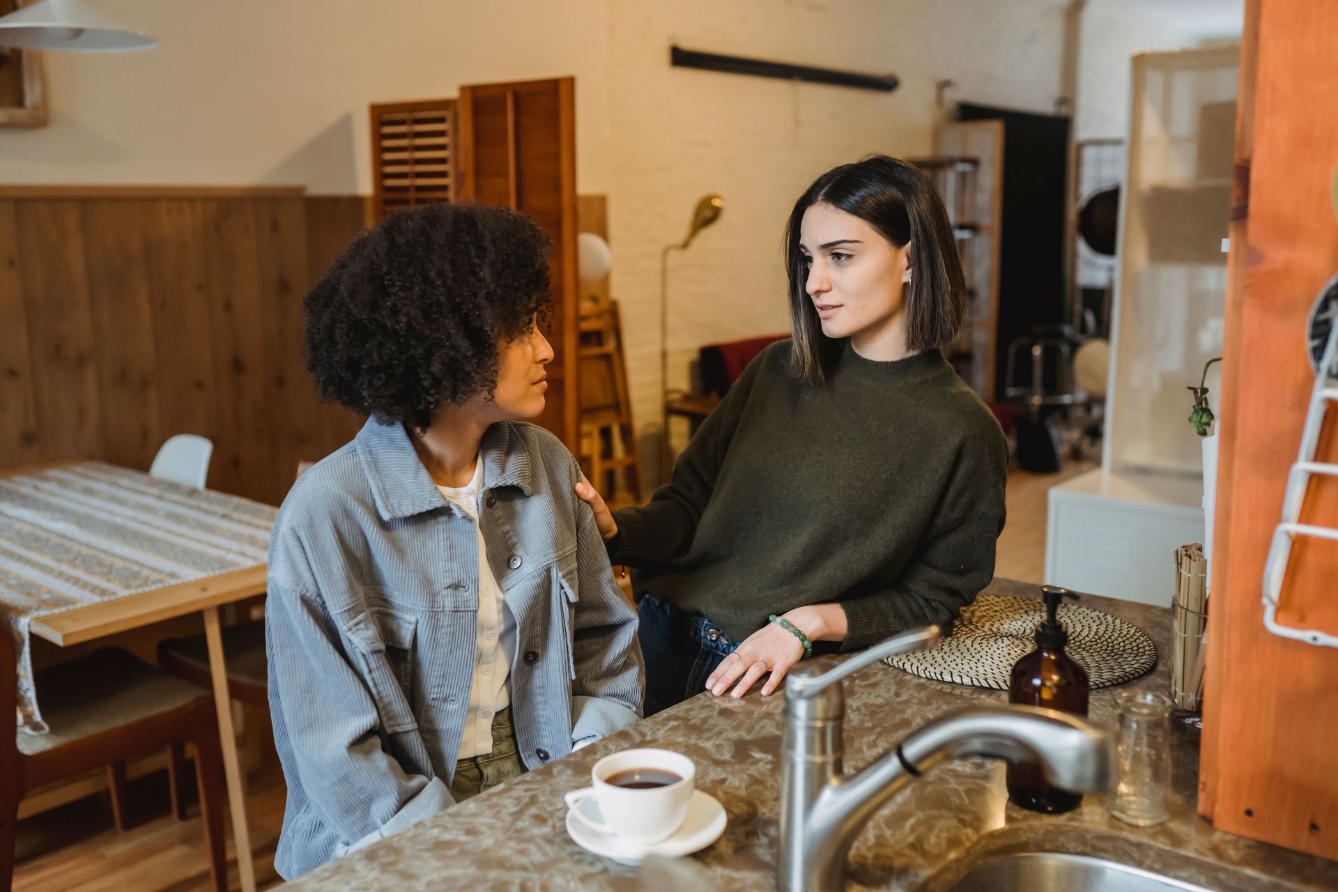 Two people talking at a kitchen counter, with cups and a sink in the foreground.