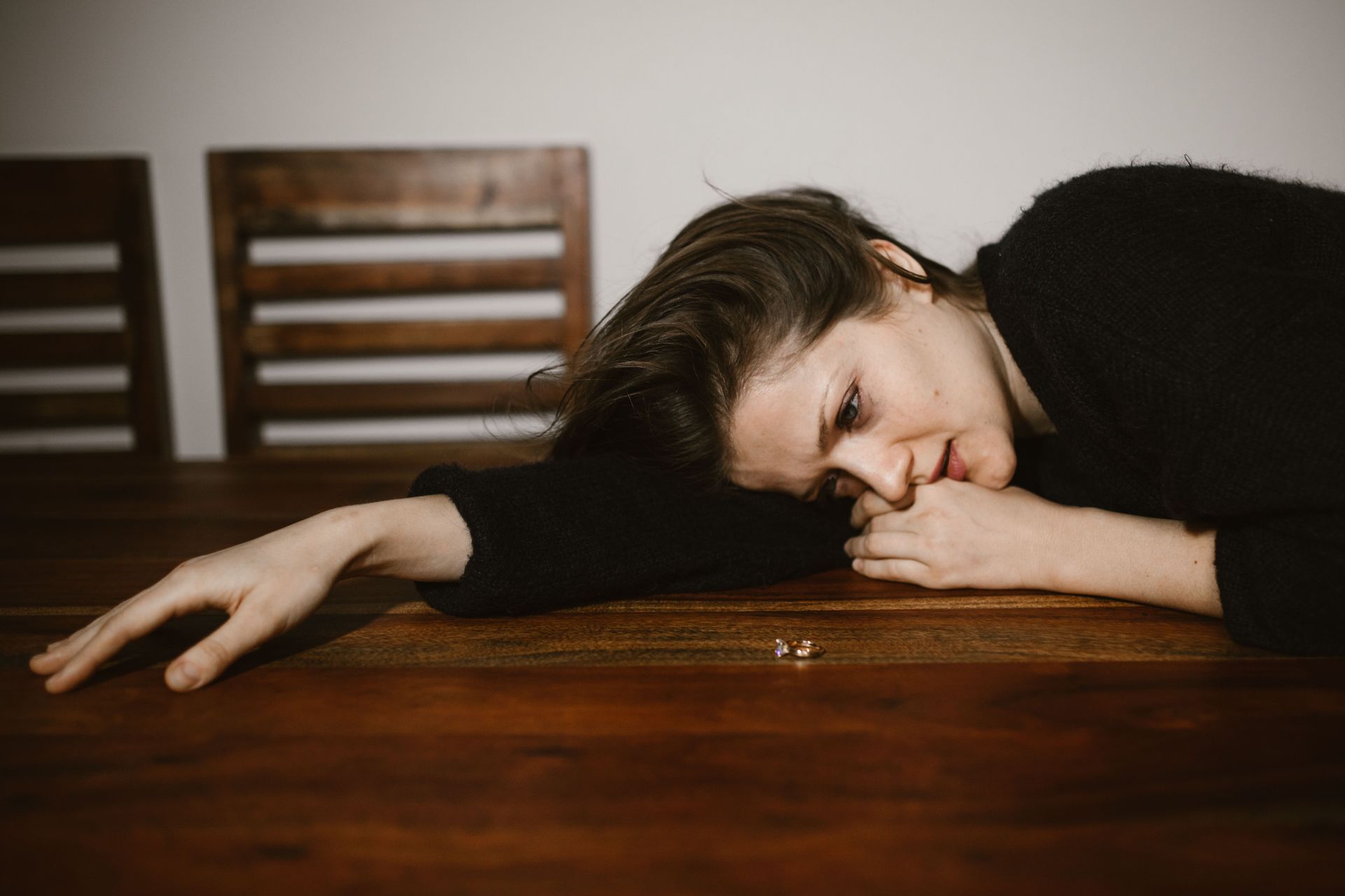 Person asleep on a wooden table, arm outstretched, in a dim room with chairs in the background
