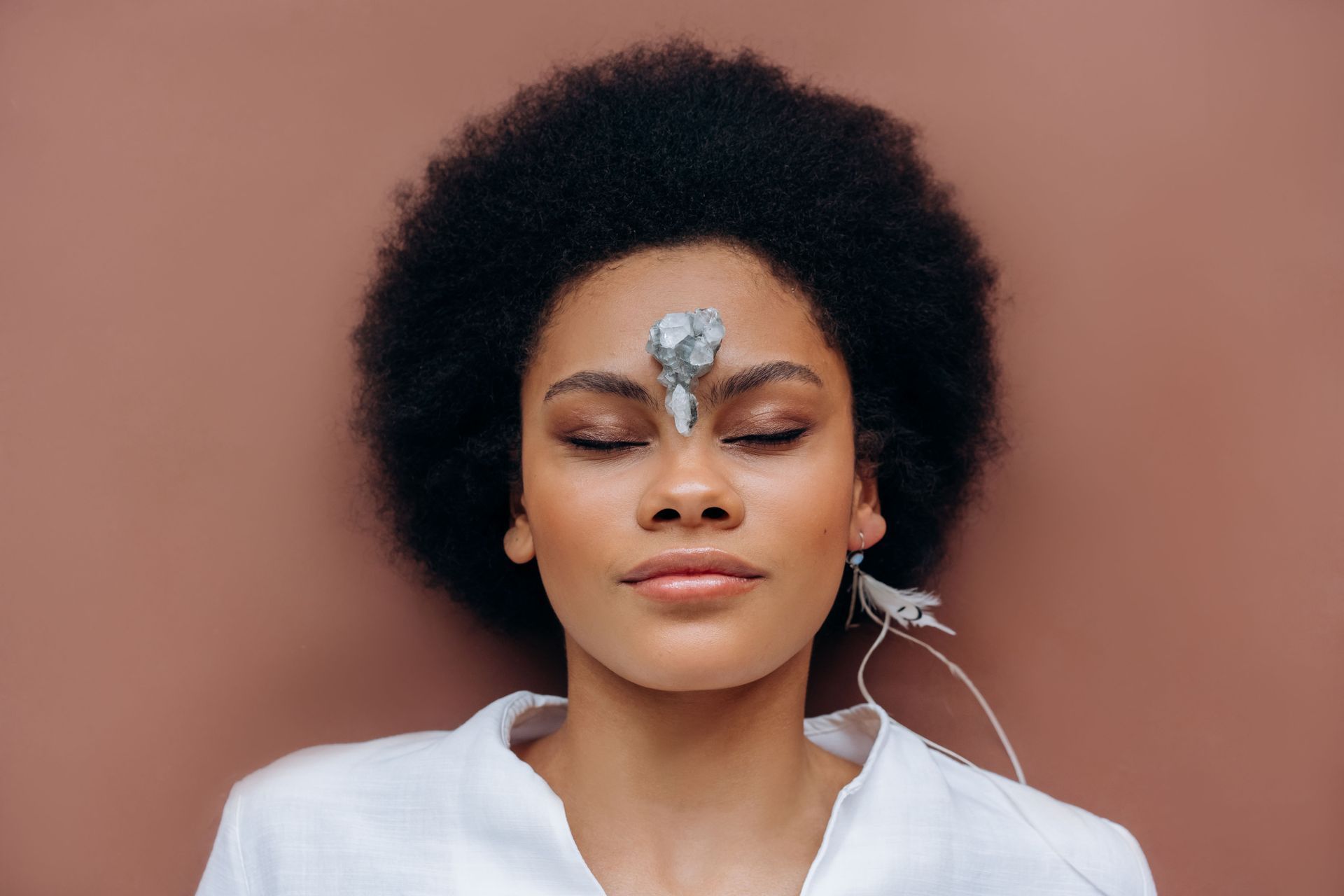 Woman with eyes closed, silver face gems, and white shirt against a pink background