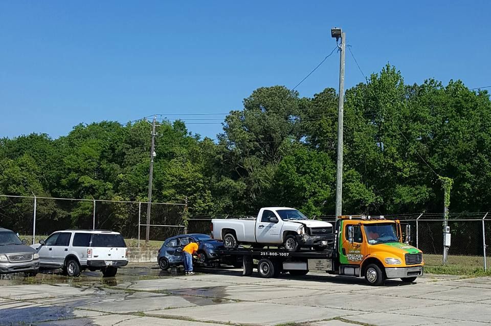 Tow truck hauling a white pickup beside a black car in a parking lot under clear blue sky