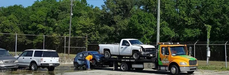 Tow truck loading a white pickup beside a blue car on a roadside with trees and a fence