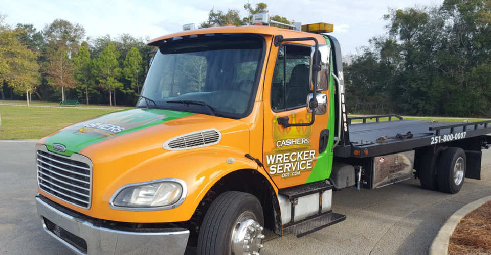 Orange and green flatbed tow truck parked on a road near a grassy field.