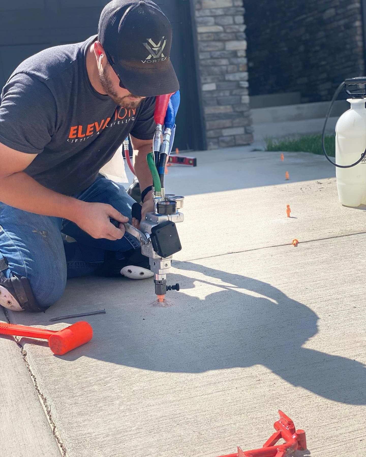 Man kneeling on concrete, using a machine. Orange flags, red hammer, and a white sprayer are nearby.