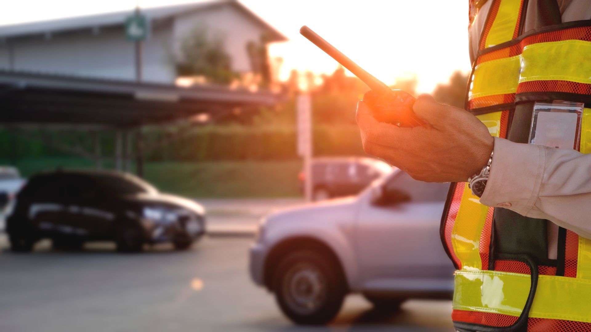 Person in high-visibility vest holding a walkie-talkie, directing traffic. Cars and buildings are in the background.