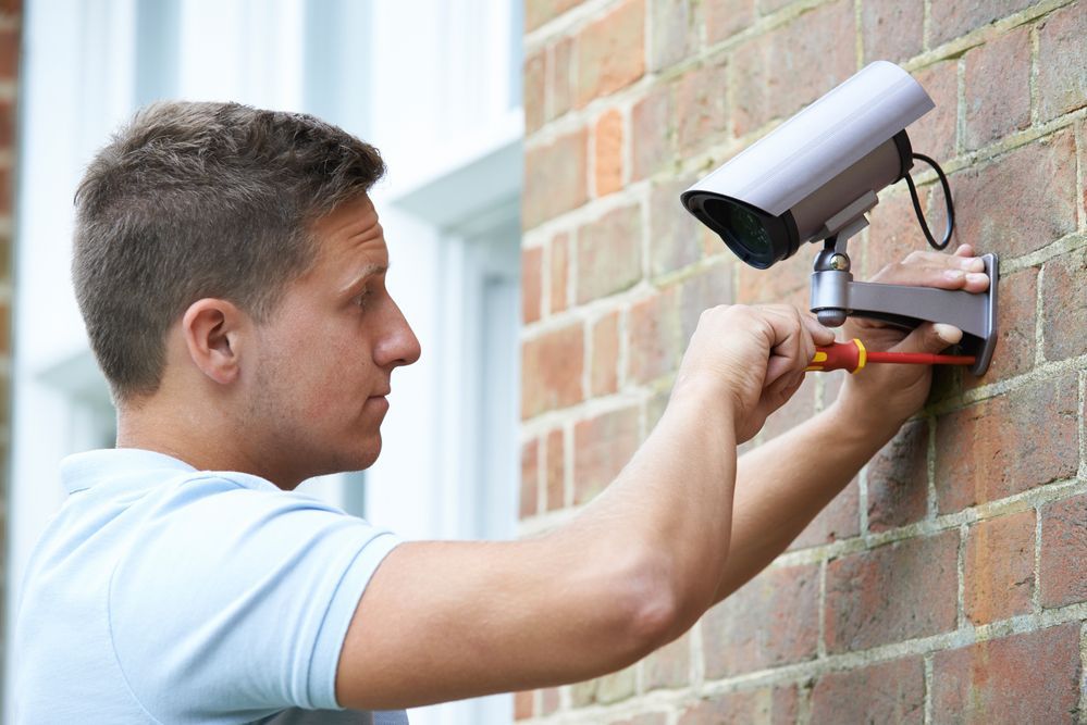 Man in light blue shirt installing security camera on a brick wall with a screwdriver.