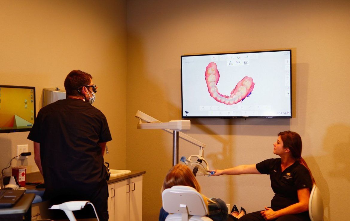 A woman is sitting in a dental chair while a man and a woman are standing in front of a television.