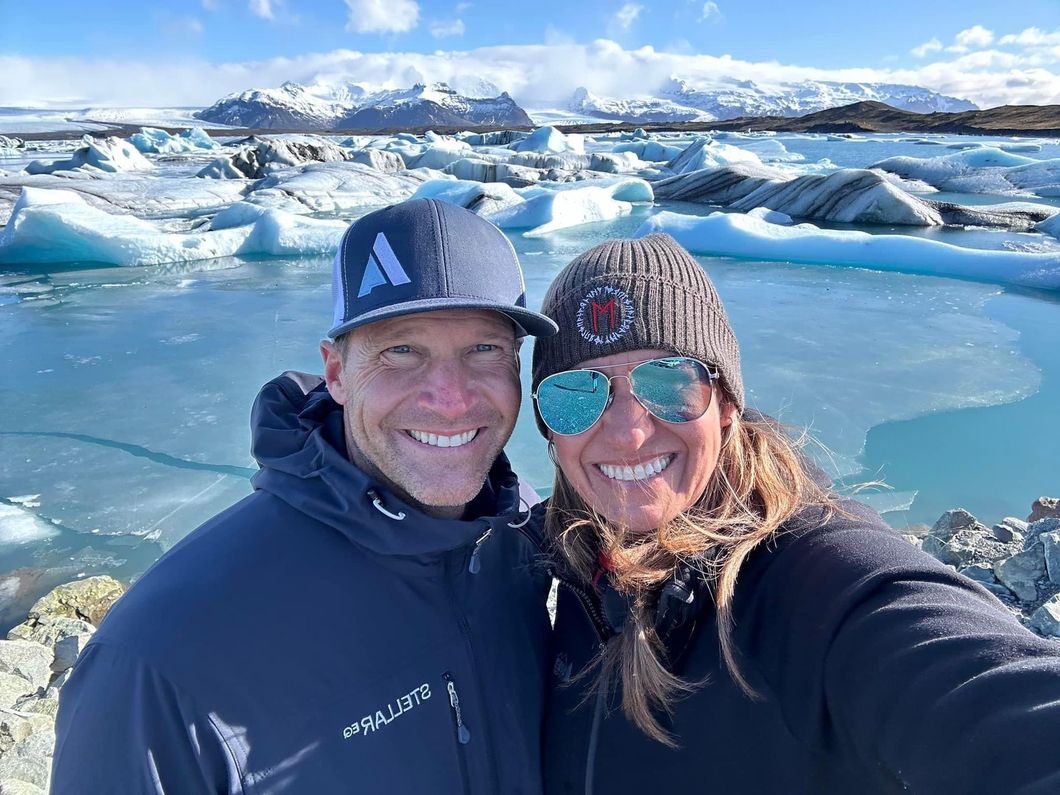 Two people smiling for a selfie by icy blue water with snow-covered mountains in the background