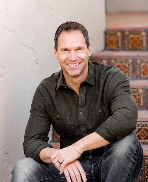 Smiling man in dark shirt and jeans sitting on steps beside a wall