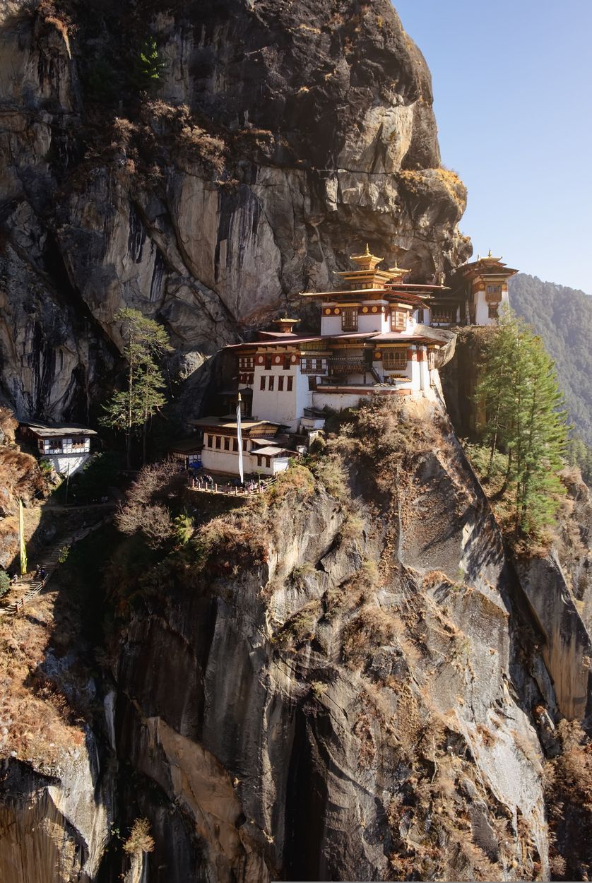 Temple clinging to a steep cliff beside a rocky mountain face, with golden roofs and blue sky.