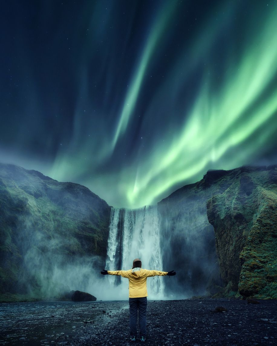 Person in a yellow jacket with arms outstretched before a waterfall under green aurora lights.