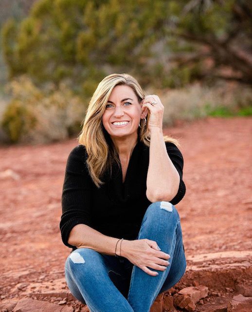 Woman in black top and jeans sitting on red rocks outdoors, smiling at the camera.