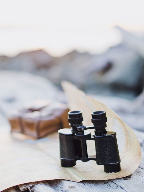 Black binoculars on a pale map or paper with a blurred backpack in the background