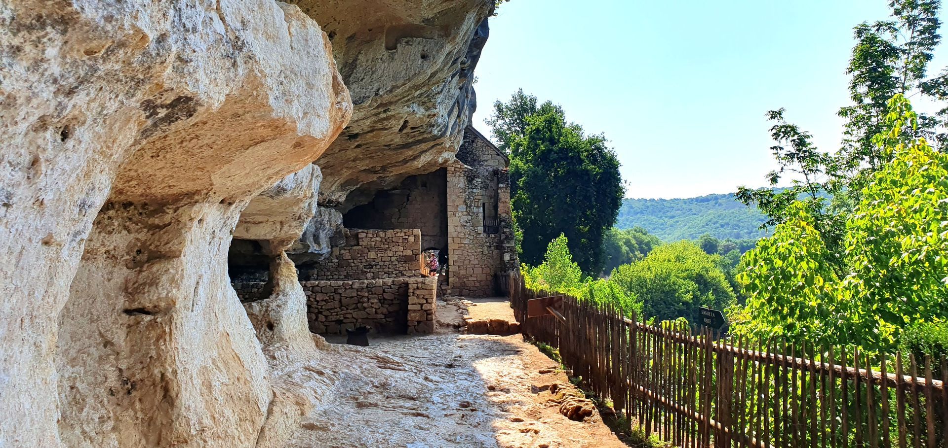 Rocky cave overlook with a shaded stone path, wooden fence, and distant green hills under blue sky