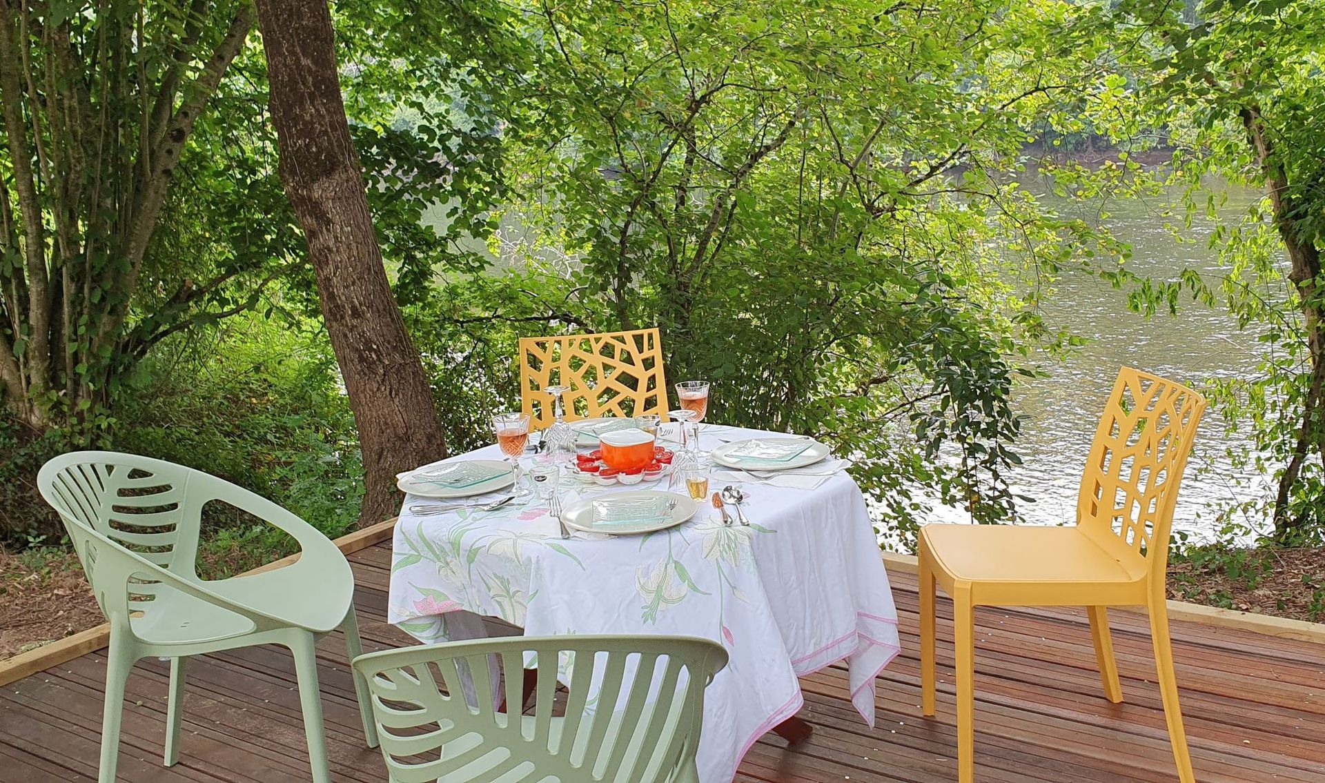 Een tafel gedekt voor een maaltijd op een houten terras aan het water, omgeven door bomen.
