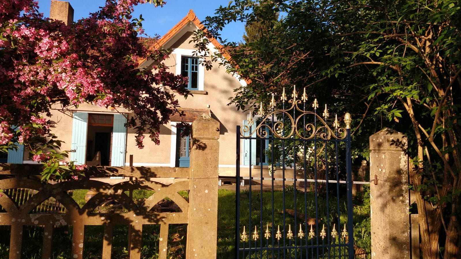 Une maison aux volets bleus et aux fleurs roses se trouve derrière un portail en fer forgé