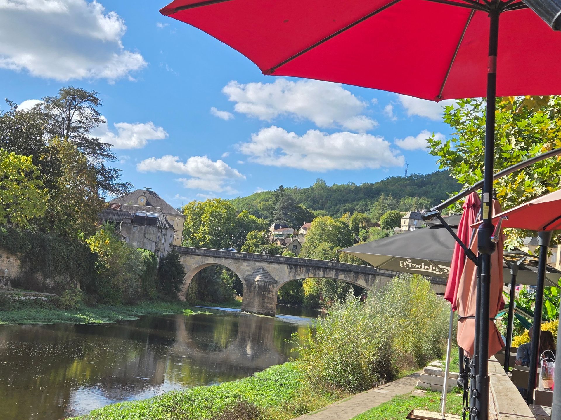 Stone bridge over a calm river with a red umbrella in the foreground and a sunny blue sky.