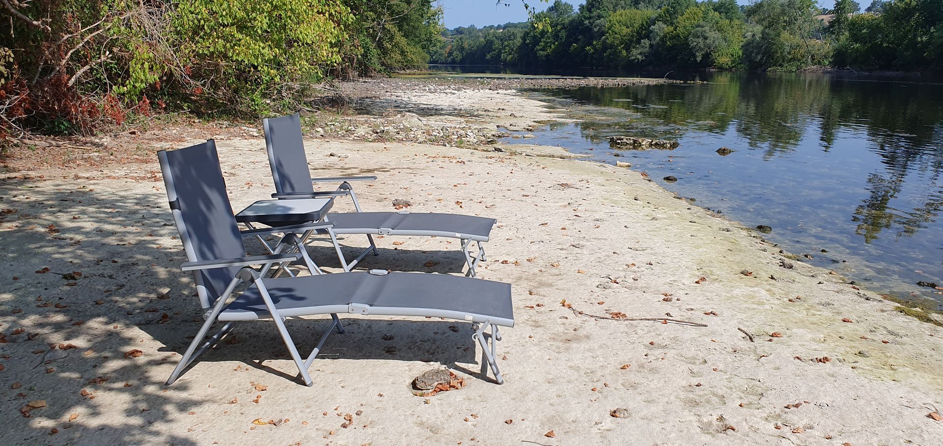 Twee klapstoelen op een zandstrand aan het water, omgeven door begroeiing.