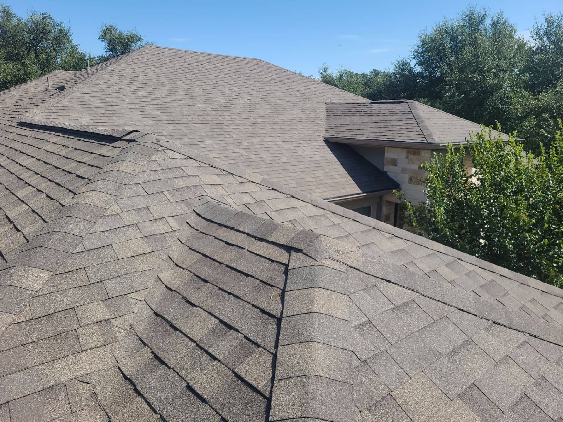 Gray asphalt shingle roof on a house, with trees in the background under a blue sky.
