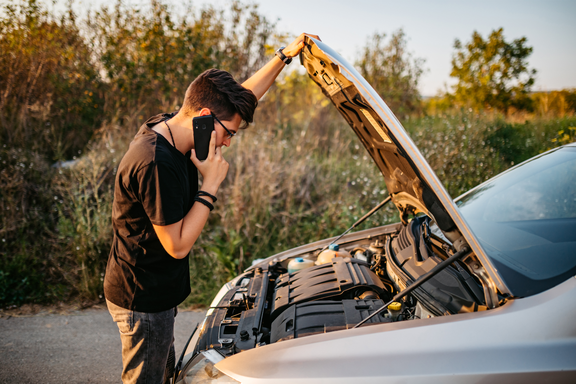 Person with phone looking into a white car’s open hood by the roadside at sunset