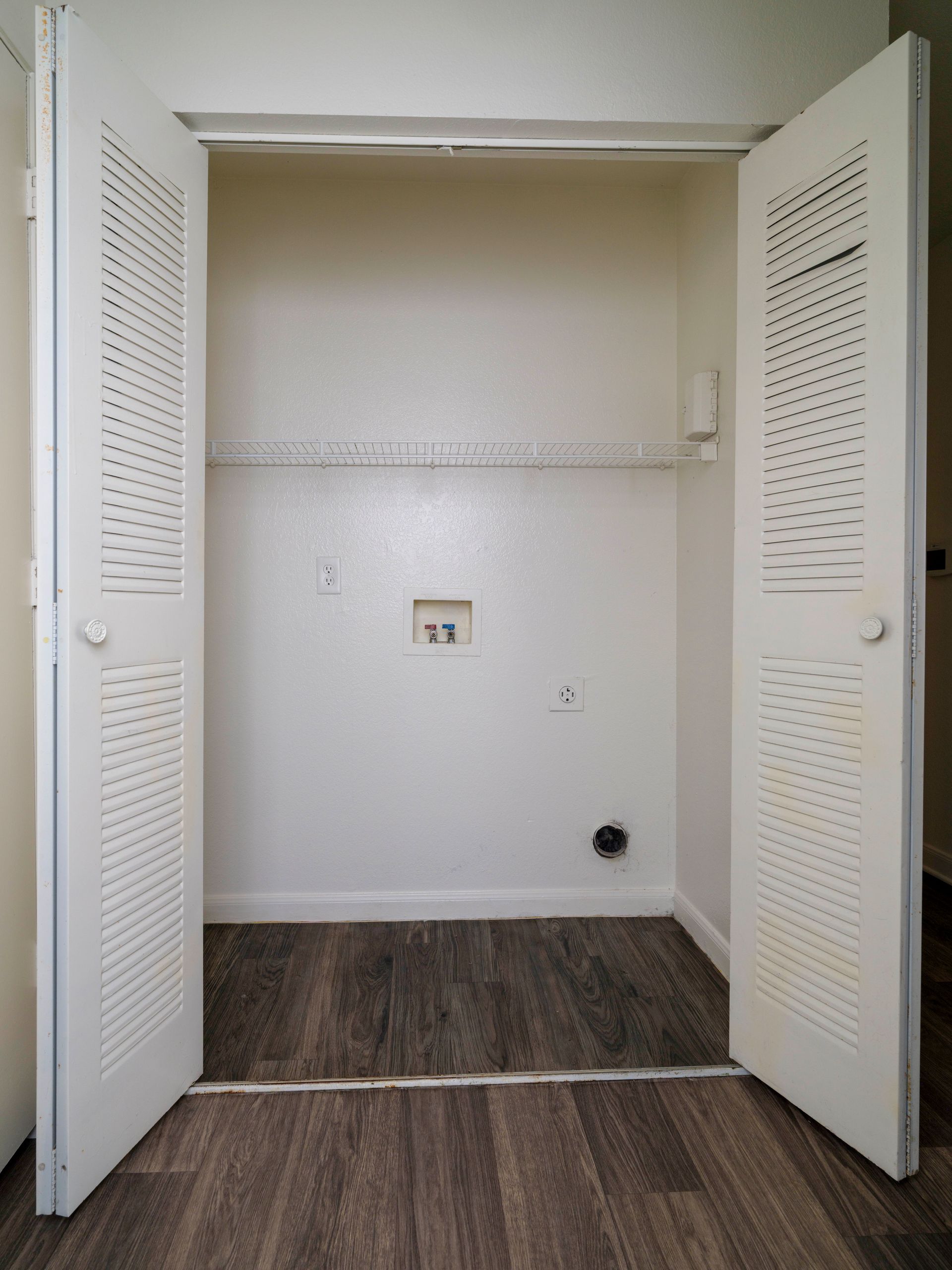 Open closet with white louvered doors and a small wall outlet on a wood-look floor.