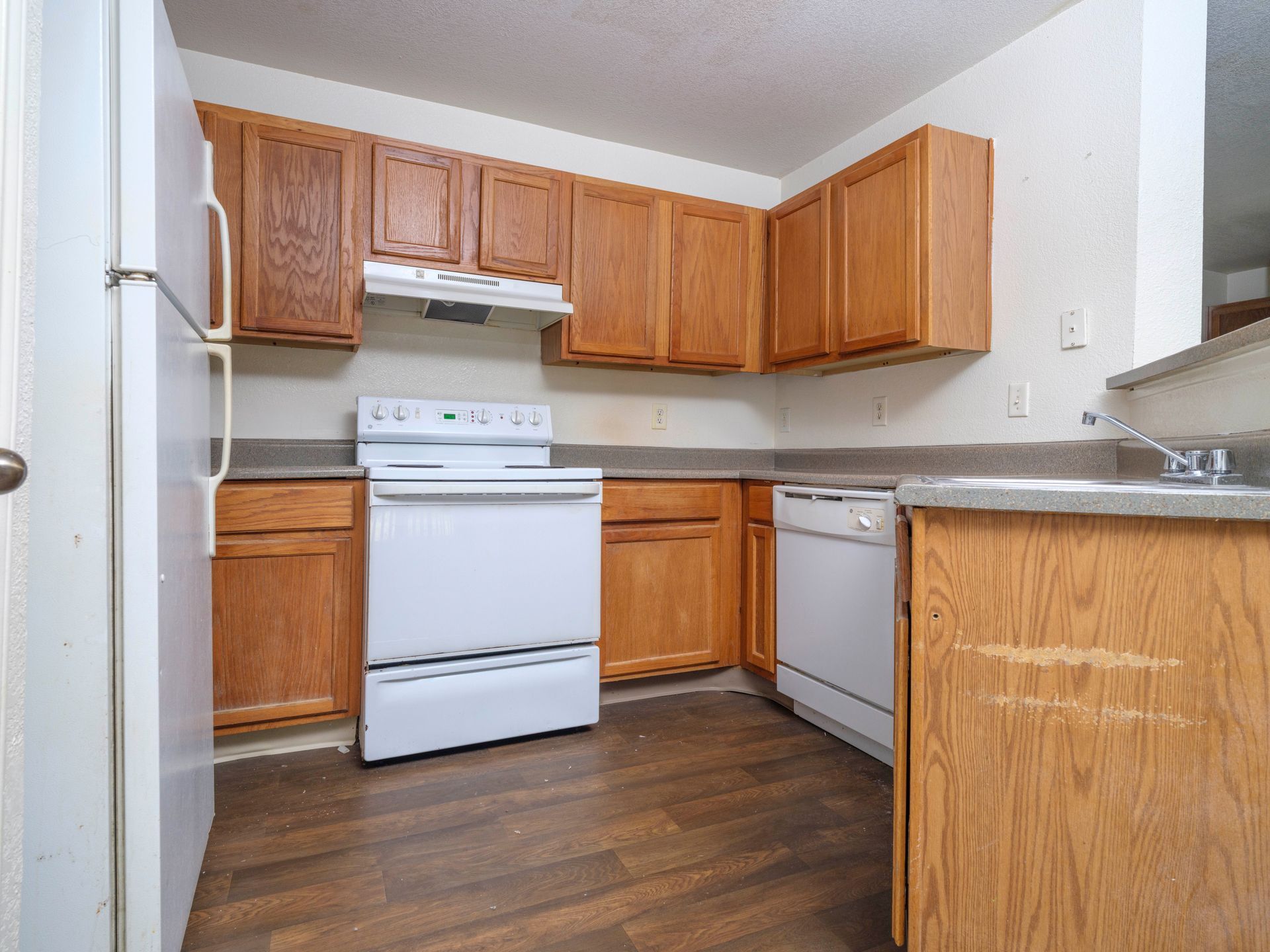 Small kitchen with wood cabinets, white stove and dishwasher, and a refrigerator on the left.
