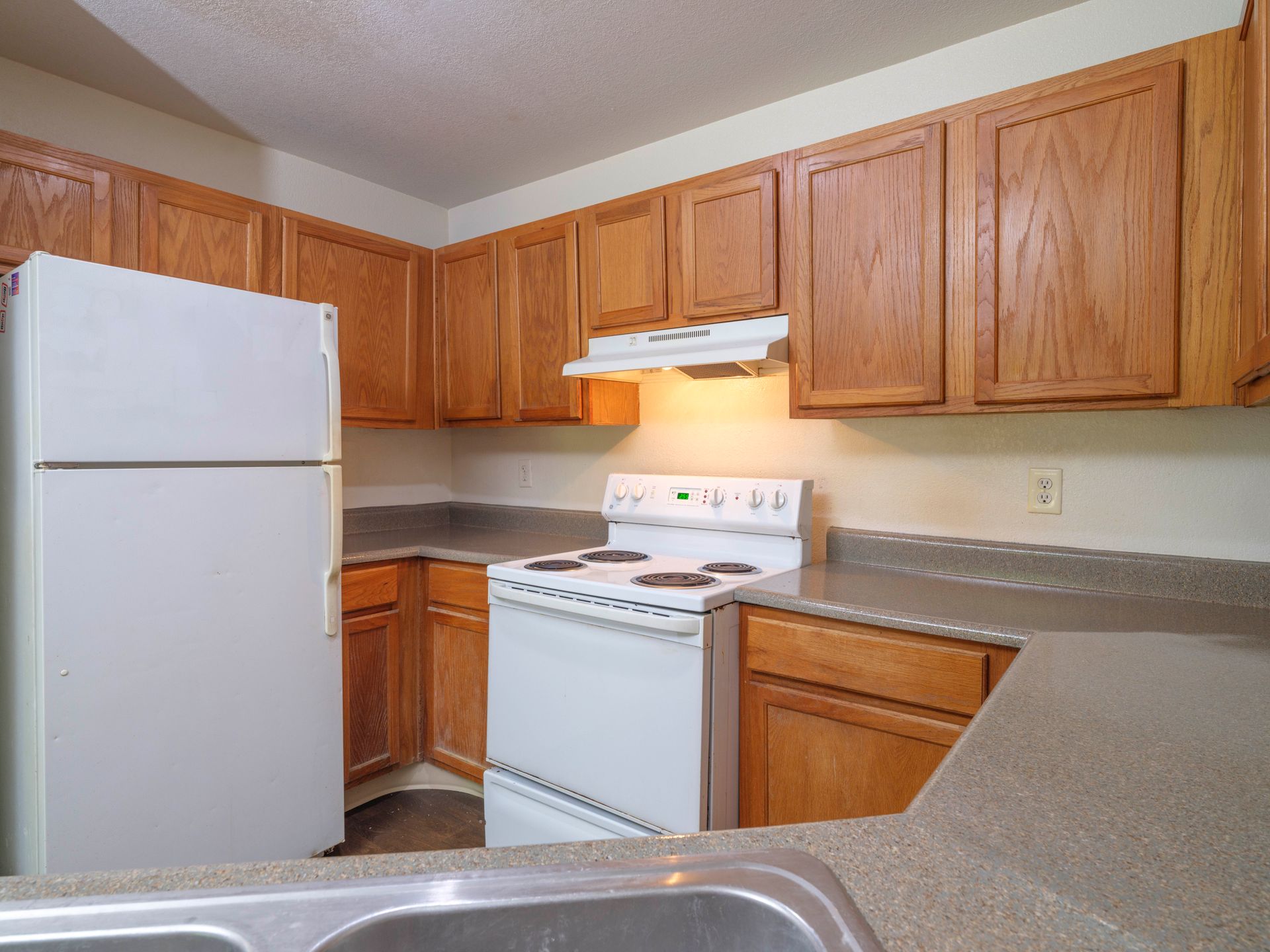 Empty kitchen with white appliances, wood cabinets, and beige countertops.