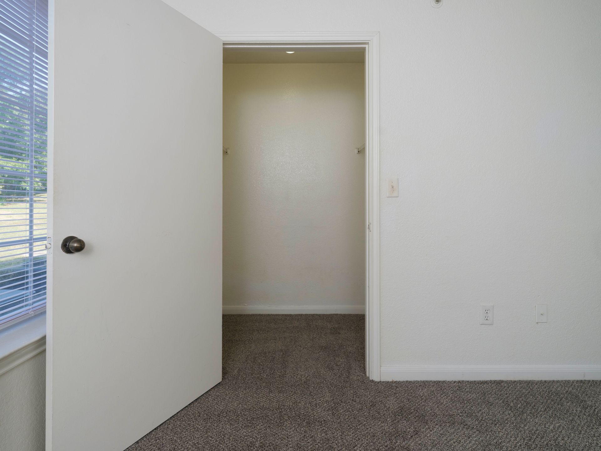 Empty room with open white door, beige walls, and carpeted floor near a window