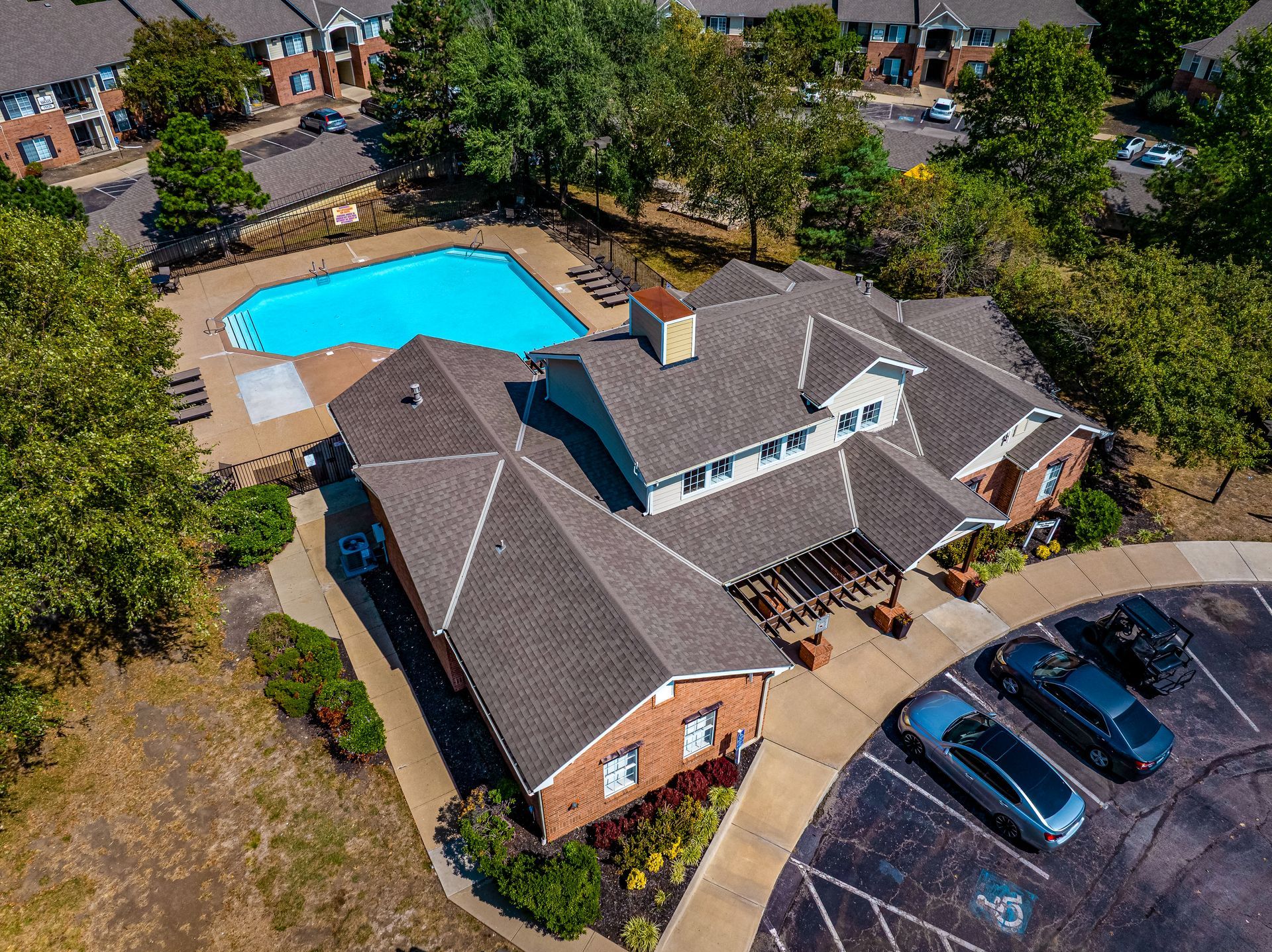 Aerial view of a house with a backyard pool, driveway, and parked cars surrounded by trees.