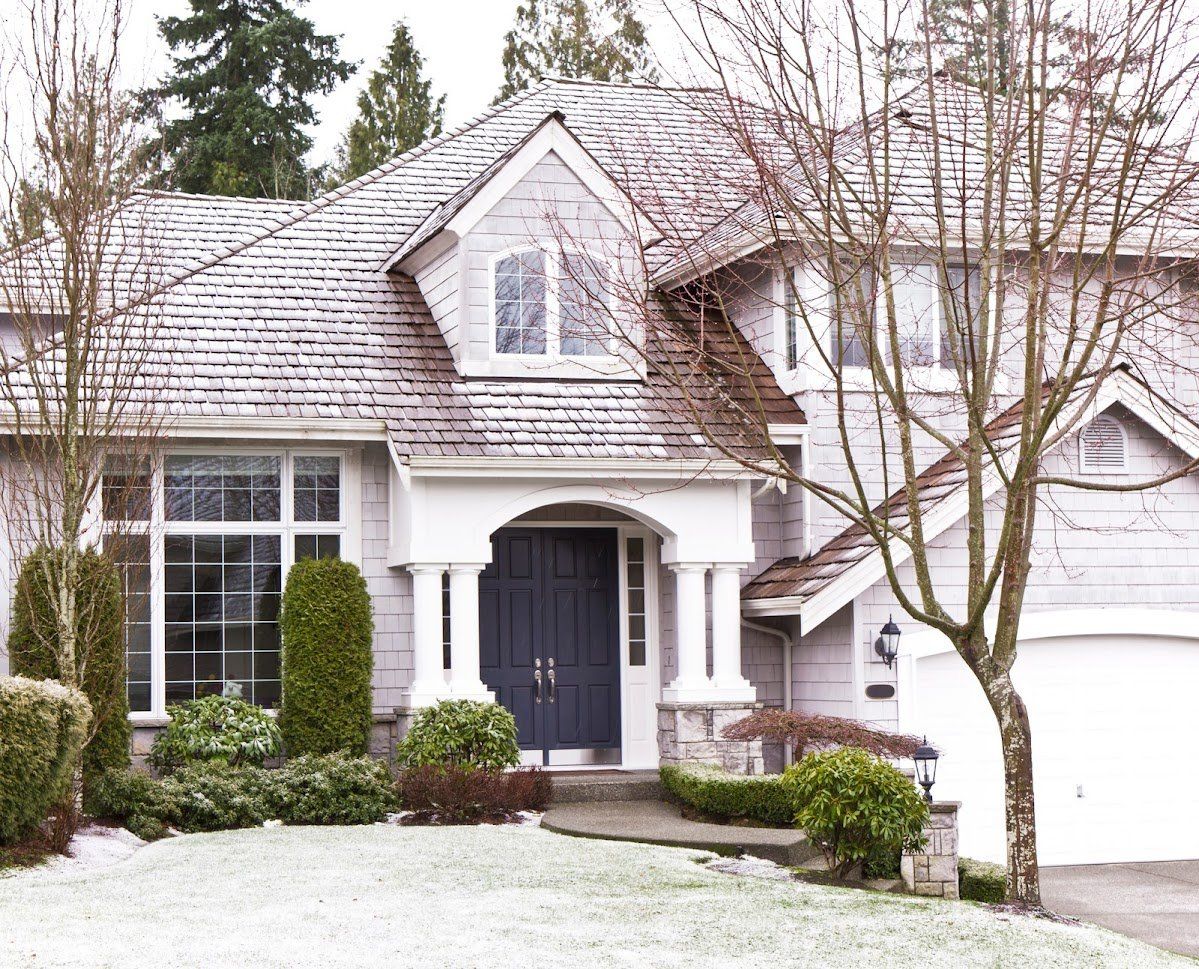 A light-colored house with a snowy roof and front yard, flanked by bare trees and green bushes.