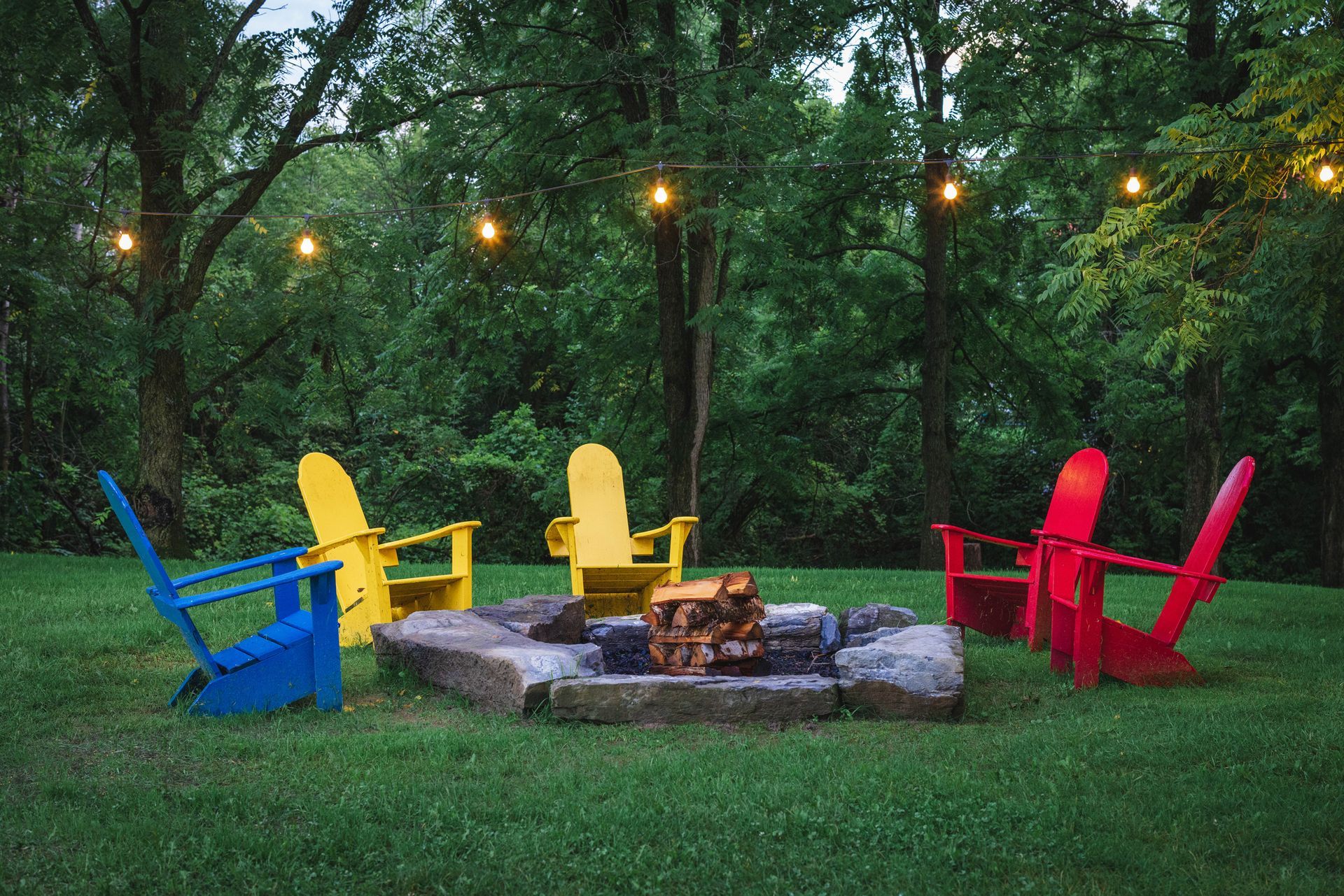 Four Adirondack chairs around a fire pit under string lights in a grassy yard, surrounded by trees.