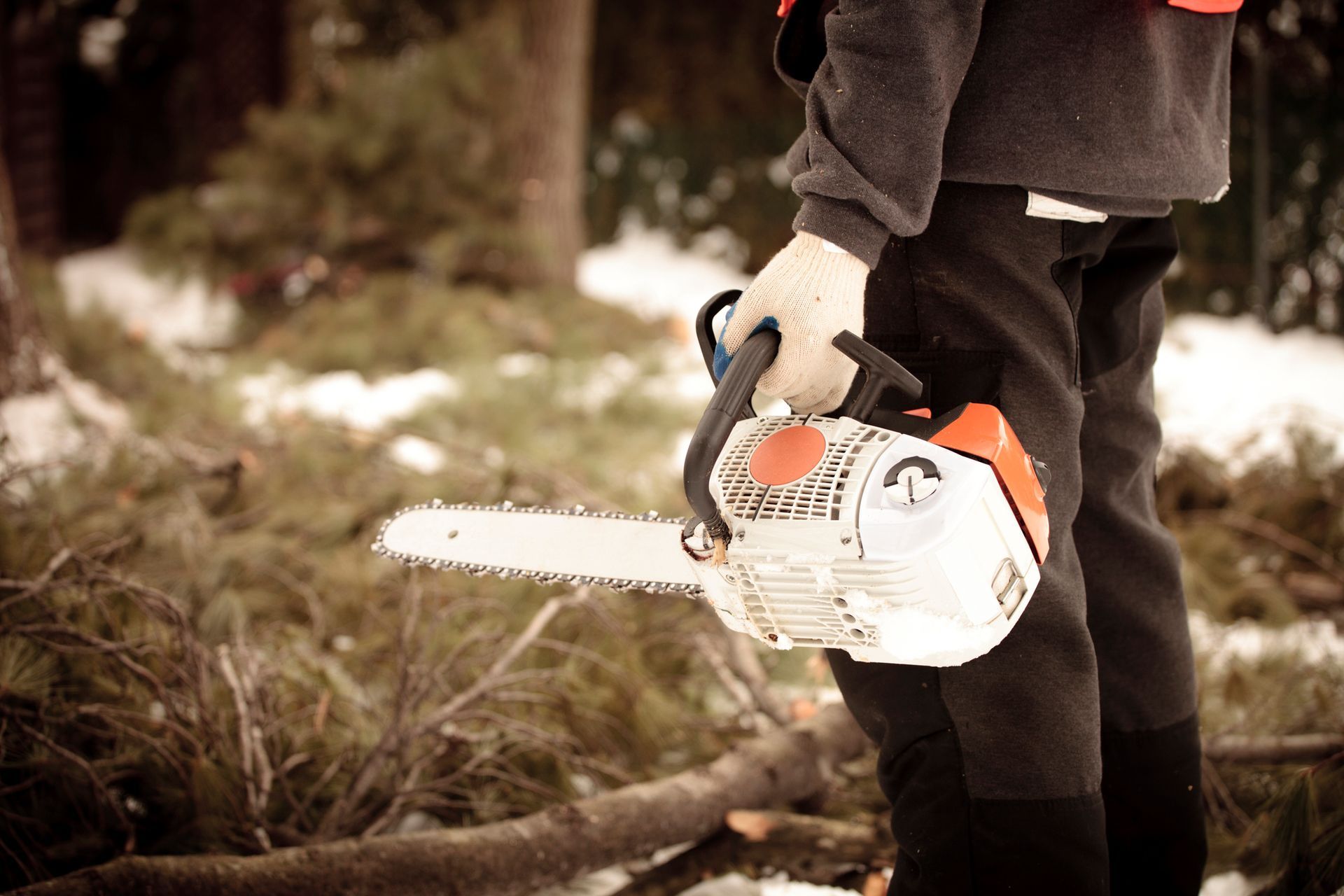 Person in work clothes holding a chainsaw outdoors, near cut tree branches and snow.