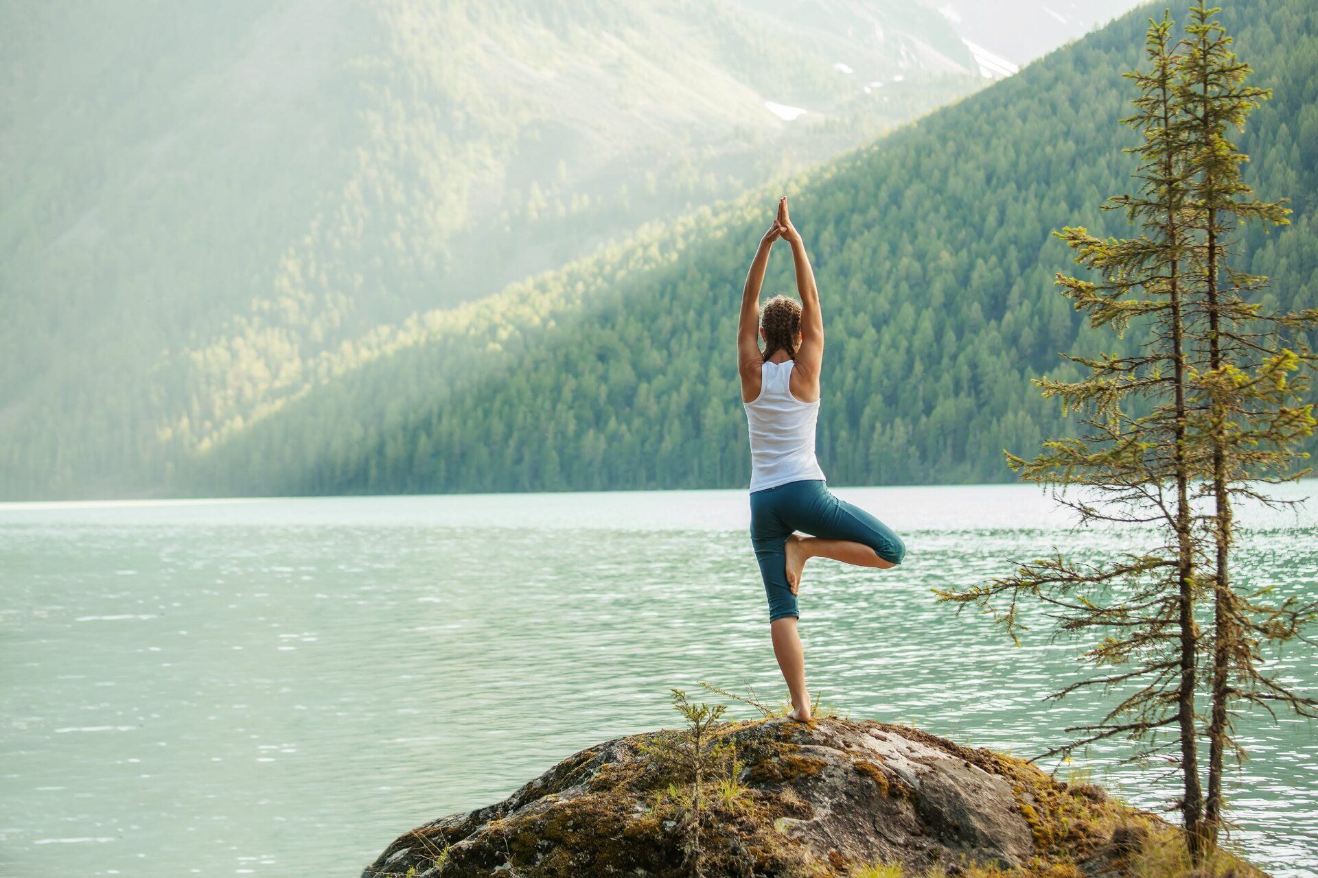 Woman in yoga pose on a rock by a lake, arms raised, green mountains in the background.