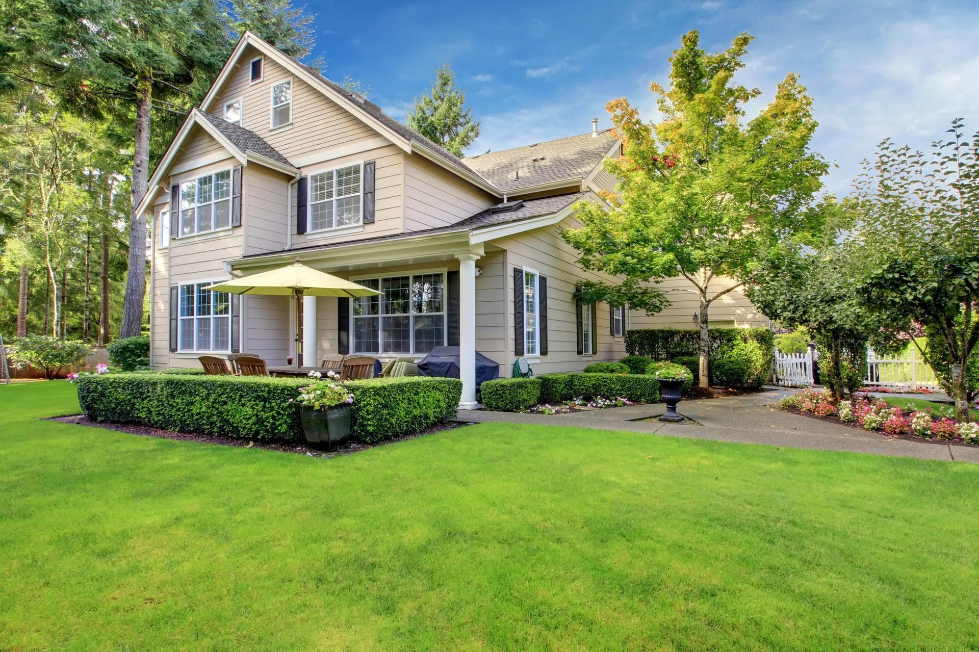 Tan house with green lawn, covered patio, shrubs, and trees under a blue sky.