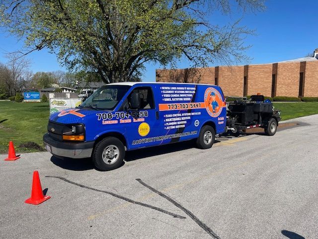 Blue utility van towing equipment beside orange cones on a street, with a brick building and tree in the background