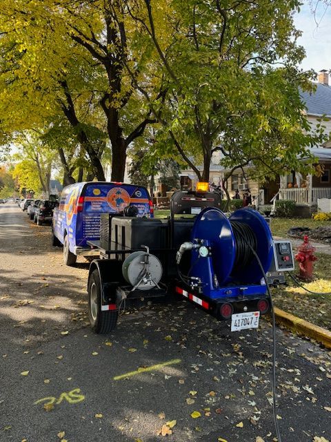 Blue utility vehicle parked on a leaf-strewn street beside a fire hydrant and another parked car