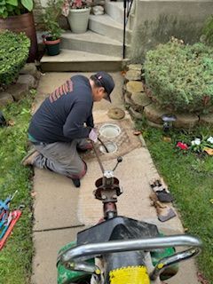 Worker kneeling by a driveway trench, repairing underground pipes beside a garden path.
