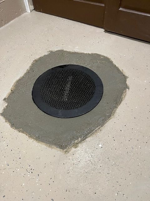 Round floor vent in a patched concrete floor near a wooden door.