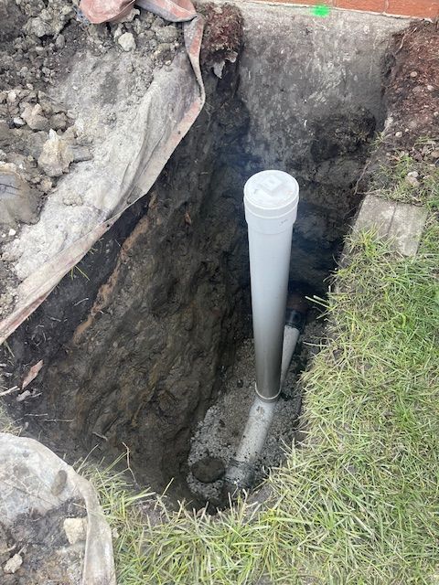 White PVC pipe standing in a deep trench beside soil, rocks, and grass