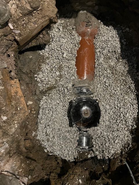Broken buried pipe valve with orange handle surrounded by gravel in a dark trench